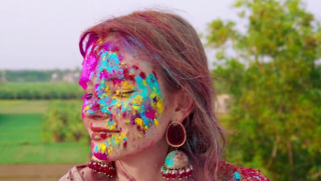 Caucasian young woman celebrating Holi face covered with multicolor paint touching hair wearing sunglasses outside. Holi festival of colors. India, traditions, jewelry, body art, artistic make up