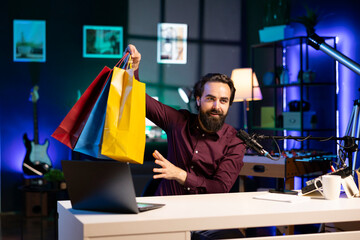 Influencer holding colorful shopping bags in hand, showing subscribers purchases he recently got. Content creator presenting fanbase new acquisitions, filming himself in apartment