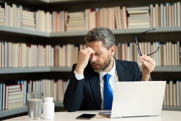 Millennial Businessman in office having headache. Business man in suit work on laptop at home office take off glasses suffer from migraine or headache. Tired exhausted man suffering from headache.