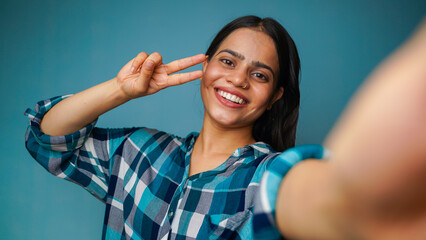 Fototapeta premium Young woman taking selfie portrait, POV shot of a beautiful Asian Indian girl taking her picture and showing peace sign or v-sign