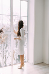 curly-haired woman in a white dress looks out the window