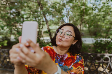 Business casual professional taking a selfie on her cell phone while enjoying nature. Adult asian,entrepreneur with digital devices capturing selfie, staying connected in an urban green space