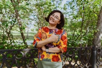 Joyful young Asian woman showing thumbs up, enjoying sunny day in the park. Happy Stylish lady in a top with a floral pattern is smiling standing at the railing of the park.