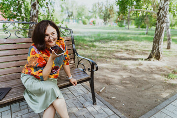 Smiling freelancer woman wearing colorful blouse, animatedly chatting on her smartphone outdoor. Entrepreneur with digital devices, passionately engaged in loud phone discussion in urban green space
