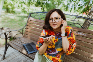 Happy woman sitting on a bench in a green park, emotionally talking on her phone. Business casual professional deeply engrossed in a phone call while peacefully seated in nature