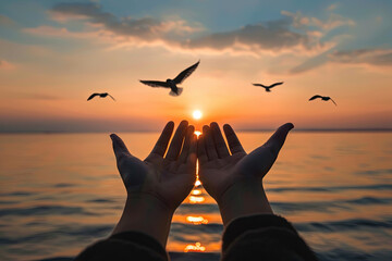 Hands outstretched upwards in a gesture of worship against a serene sunset over water, with birds soaring above. Symbolizing prayer and seeking divine blessings - obrazy, fototapety, plakaty