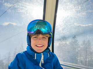 Cheerful boy in ski outfit inside lift cabin among snow peaks
