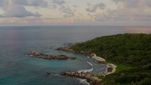 Birds-eye view of the stunning pink sunset over the granitic island. Aerial shot revealing the transparent turquoise waters, lush green vegetation, sandy beaches, and granite boulders in La Digue.