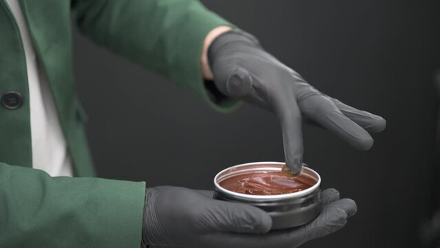 A barber in black gloves takes some pomade wax from a tin can to style a clients hair.