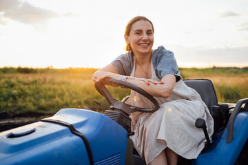 A young smiling farmer sits at the wheel of a tractor. A funny charming woman in casual clothes...