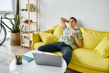 weary guy with laptop putting hand behind head and leaning on yellow couch with glasses