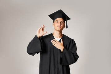 charming young student in graduate gown and cap making wish against grey background