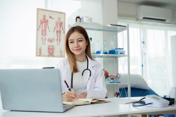 Young Asian doctor in white medical uniform with stethoscope using laptop computer video chatting with patient at table in health clinic or hospital