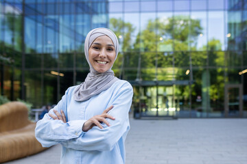 A professional woman in a hijab stands confidently with crossed arms outside a contemporary office...