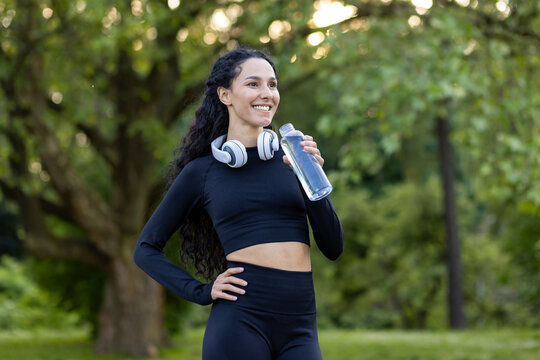 Smiling active woman in workout gear taking a break with a water bottle in a green park setting, headphones around her neck.