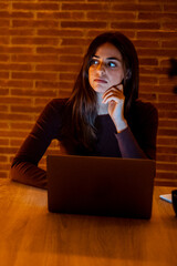Beautiful Authentic Young Woman Sitting at a Table in Cozy Kitchen and Using Laptop Computer at Home at Night. Female Doubts and Chatting on the Internet and Social Media, Doing Online Research.