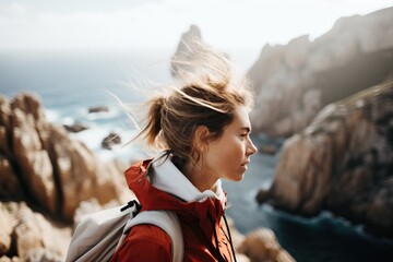 Woman with backpack stands on a cliff overlooking the ocean, wind in her hair - Powered by Adobe