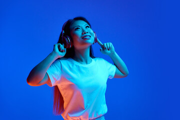 Portrait of young attractive Asian woman listening her favorite music in headphones against blue...