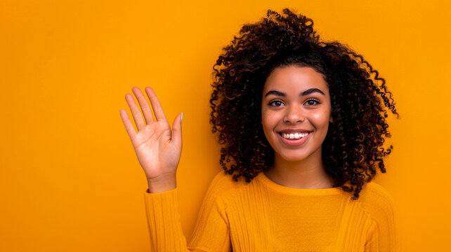 Young woman smiles and raises her hand in greeting on a yellow background.