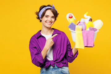 Young smiling woman wear purple shirt hold point index finger on basin with detergent bottles do...