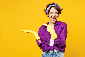 Young cheerful woman wear purple shirt rubber gloves while doing housework tidy up point index finger on empty palm with area isolated on plain yellow background studio portrait. Housekeeping concept. - obrazy, fototapety, plakaty