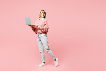 Full body elderly IT woman 50s years old wears sweater shirt casual clothes glasses hold use work on laptop pc computer look aside isolated on plain pastel pink background studio. Lifestyle concept.