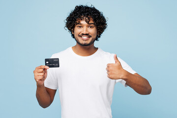 Young happy smiling Indian man wear white t-shirt casual clothes hold in hand mock up of credit bank card show thumb up isolated on plain pastel light blue cyan background studio. Lifestyle concept