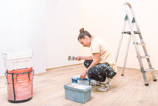 A focused woman preps her paintbrush, surrounded by paint cans and a ladder, signaling the beginning of a home painting project
