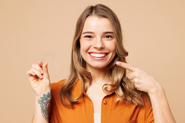 Young happy woman she wear orange shirt casual clothes hold in hand invisible transparent aligners, invisalign bracer point on teeth isolated on plain pastel light beige background. Lifestyle concept.