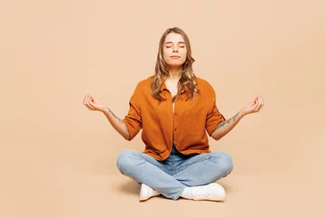 Tuinposter Full body young woman wear orange shirt casual clothes sits hold spread hands in yoga om aum gesture relax meditate try to calm down isolated on plain pastel light beige background. Lifestyle concept. © ViDi Studio