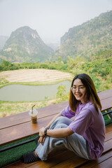 Fototapeta na wymiar Asian woman drinking coffee and solo travel on tropical mountain peak in summer sunny day. Attractive girl enjoy and fun outdoor lifestyle looking camera with moutain view background.