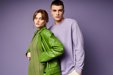 fashionable couple in vibrant clothes posing together on purple background and looking at camera