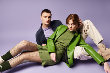 fashionable couple in vibrant attires sitting on floor and looking at camera on purple backdrop