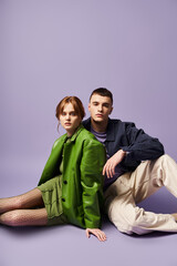 Obraz premium fashionable couple in vibrant attires sitting on floor and looking at camera on purple backdrop