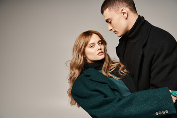 appealing man in coat posing lovingly next to his beautiful girlfriend who looking at camera