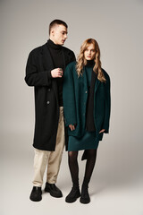 good looking stylish boyfriend and girlfriend in coats posing lovingly together on gray backdrop