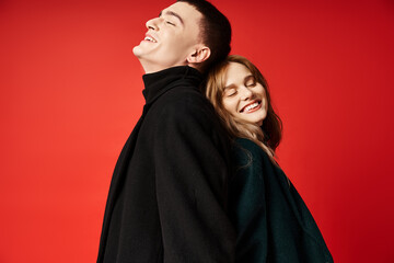 cheerful loving couple in stylish coats smiling happily with closed eyes on red vivid backdrop