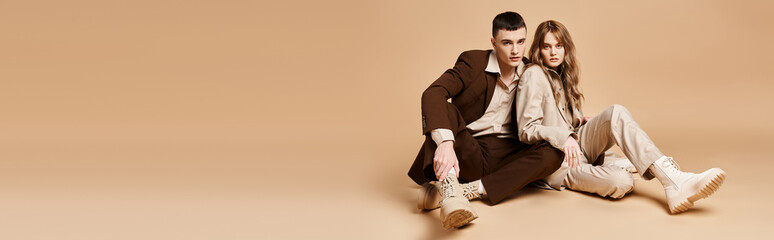 appealing couple in elegant suits sitting on floor and looking at camera on pastel backdrop, banner