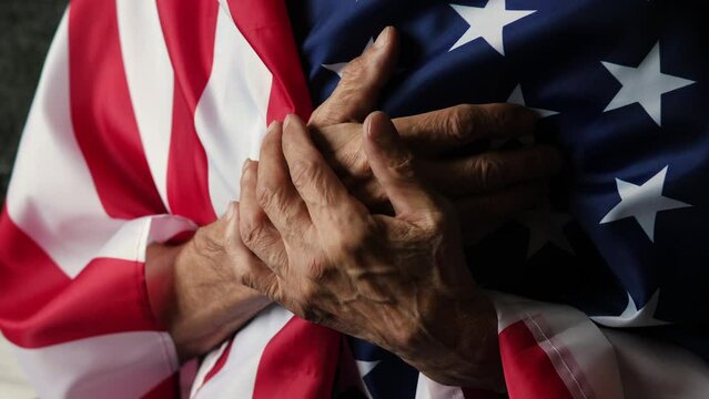 an elderly American man hugging the American flag listens to the country's national anthem while holding his heart. patriotic feelings of a pensioner