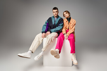 loving appealing couple in vivid stylish bombers looking at camera on gray backdrop on white cube