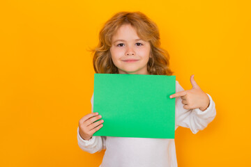 Fototapeta na wymiar Smiling kid showing index finger on green empty sheet of paper, isolated on yellow background. Portrait of a kid holding a blank placard, poster.
