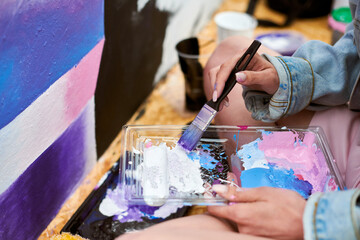 Fototapeta premium Female painter hand dips paintbrush into palette of colorful paints for live painting of picture for outdoor street exhibition, close up view of female artist hand holding paintbrush