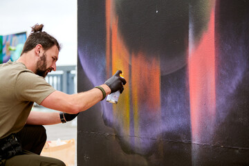 Attractive male artist is painting picture with paint spray can spraying it onto canvas at outdoor...
