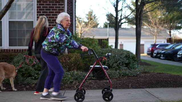 Side view of elderly senior woman pushing a walker with daughter and dog besides her, rolling on a sidewalk outside for exercise and mobility.