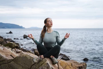 Poster Young spiritual calm happy woman with closed eyes meditate in lotus pose at the sea, om aum gesture, girl relaxing outdoors, meditation, try to calm down in the mountains, female doing yoga asana © Соня Монштейн