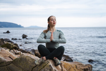 Young calm smiling woman looking forward and praying in lotus pose at the sea, girl relaxing outdoors, meditation, try to calm down, female in sport clothes keeping palms together, please God help me