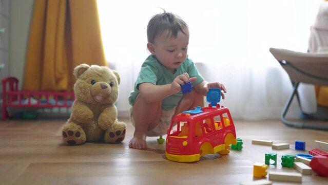 baby boy plays with toy bear a toy bus and puts wooden sticks in the hatch. development of fine motor skills concept. baby learns to put sticks toys into a bus car indoors in dream kindergarten