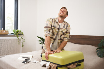 Portrait of young man sweating, shutting his suitcase, trying to close the luggage with lots of...