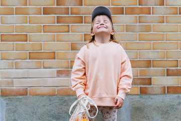 Happy little girl holding oranges in mesh bag by brick wall.