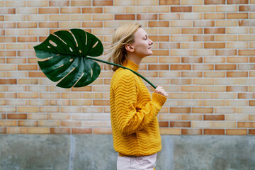 Smiling woman holding monstera leaf in front of brick wall. Concept of Ecology, Sustainability.
